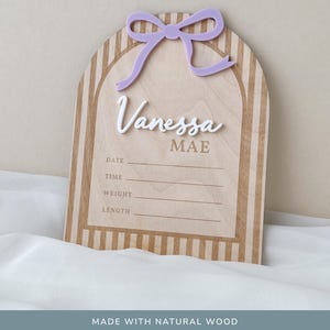 May include: An arched wooden birth announcement sign featuring a light purple acrylic bow at the top. The sign displays the name Vanessa Mae in white and brown lettering, with engraved lines for date, time, weight, and length information.