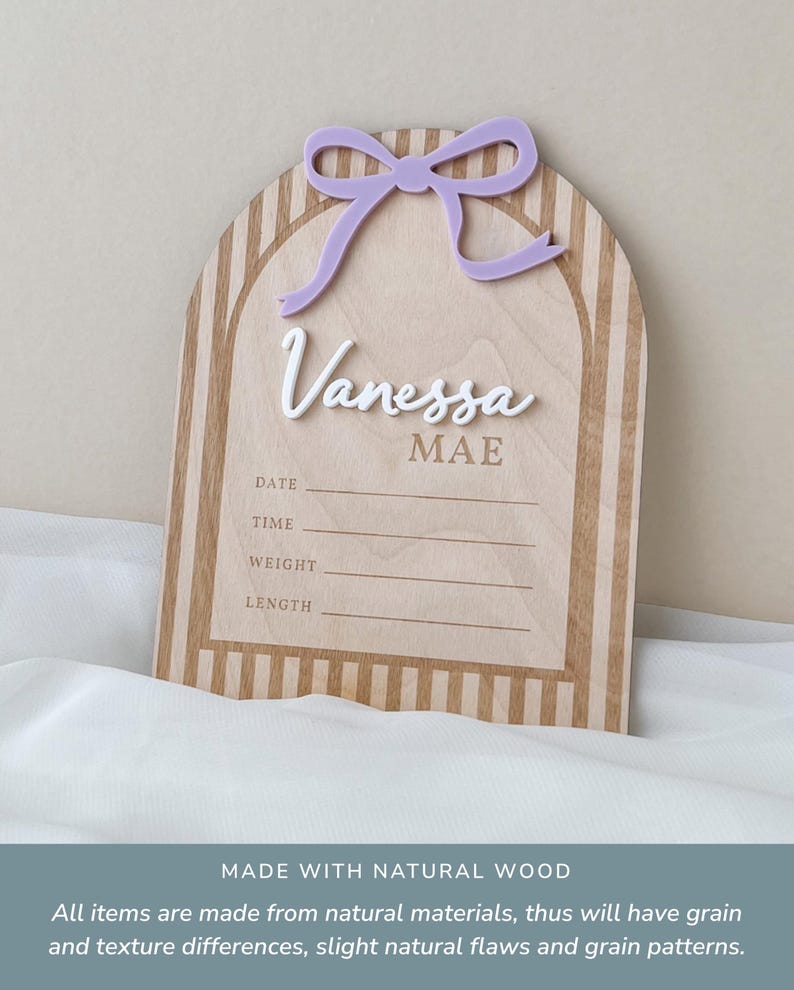 May include: An arched wooden birth announcement sign featuring a light purple acrylic bow at the top. The sign displays the name Vanessa Mae in white and brown lettering, with engraved lines for date, time, weight, and length information.