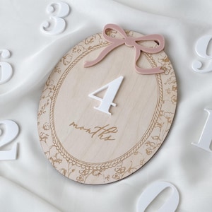 Pu&ograve; includere: Una targa ovale in legno con finitura a venature chiare e bordo floreale dorato. La targa presenta un numero 4 bianco in rilievo, la parola months in corsivo e un fiocco in acrilico rosa polvere fissato al centro della parte superiore.