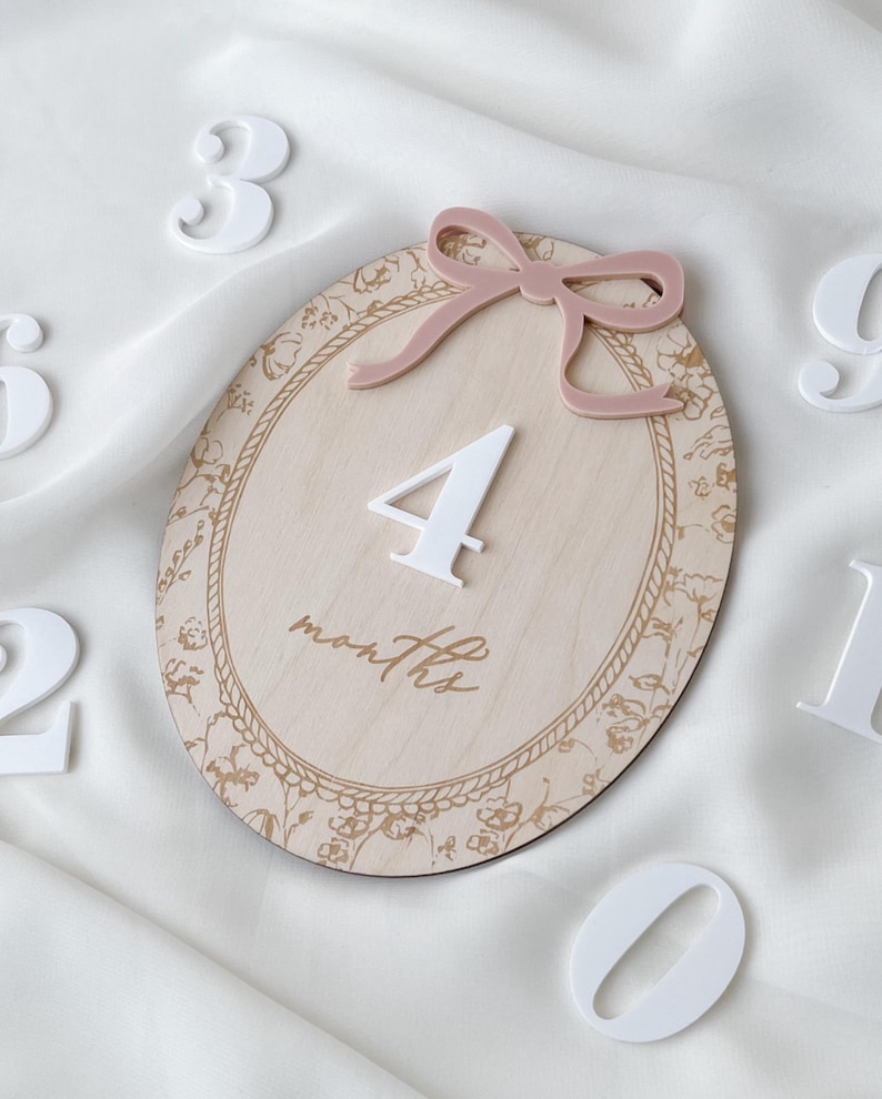 Pu&ograve; includere: Una targa ovale in legno con finitura a venature chiare e bordo floreale dorato. La targa presenta un numero 4 bianco in rilievo, la parola months in corsivo e un fiocco in acrilico rosa polvere fissato al centro della parte superiore.