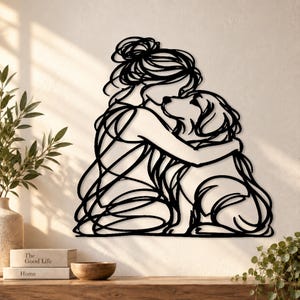 Puede incluir: Una pieza de arte mural de metal negro con un dibujo de línea continua de una persona con el cabello recogido en un moño abrazando a un perro. El diseño está compuesto por líneas negras fluidas e interconectadas que forman las siluetas.
