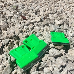 Könnte beinhalten: Drei grüne Kunststoff-Schaber mit Metallklingen. Die Schaber liegen auf einem Bett aus kleinen grauen Steinen.