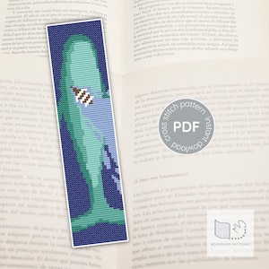Puede incluir: Un marcapáginas con un diseño de punto de cruz que presenta una ballena en tonos azules y verdes. El marcapáginas tiene un borde blanco y está colocado sobre el fondo de un libro abierto. El diseño incluye una pequeña abeja.