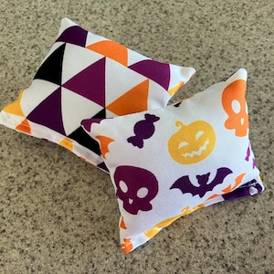 May include: Two Halloween-themed pillow covers. One features a geometric pattern in black, white, purple, and orange. The other features a white background with purple skulls, orange pumpkins, bats, and candy corn.