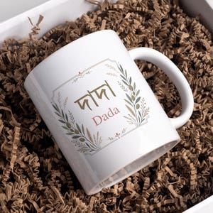 Könnte beinhalten: Ein weißer Keramikbecher mit einer goldenen bengalischen Schrift für Dada über dem englischen Text Dada in einer zartrosa Schriftart. Das Design ist von einem zarten botanischen Kranz mit grünen Blättern und kleinen braunen Akzenten umrahmt.
