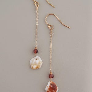 May include: A pair of gold-toned drop earrings featuring thin chains attached to curved ear wires. Each earring is accented with a small, translucent dark red bead and a single, irregular, white and brown patterned shell charm at the base.