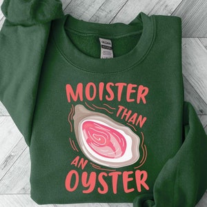 Peut inclure: Un sweat-shirt vert foncé avec le texte "MOISTER THAN AN OYSTER" en lettres roses. Le texte est accompagné d'un graphique représentant une coquille d'huître rose.