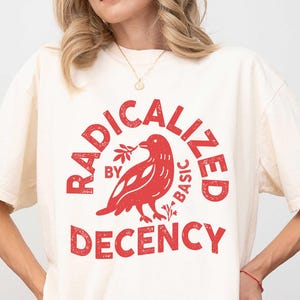 Peut inclure: T-shirt ivoire avec un motif rouge. Le motif représente un oiseau tenant une branche et les mots "RADICALIZED BY BASIC DECENCY" dans un cercle. Le t-shirt a des manches courtes et une coupe ample.