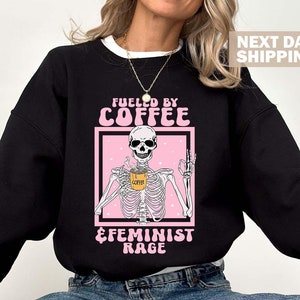 May include: Black sweatshirt with a pink graphic of a skeleton holding a coffee cup. The graphic says "Fueled by Coffee & Feminist Rage".