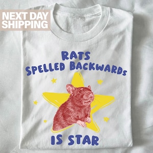 May include: White t-shirt with a graphic of a brown rat inside a yellow star. The text reads "RATS SPELLED BACKWARDS IS STAR" in blue and white. The shirt is folded neatly.