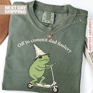 Op de afbeelding: Mosgroen t-shirt met de tekst "Off to commit dad foolery" en een cartoon kikker op een scooter, met een witte hoed. Het shirt heeft een Comfort Colors label.