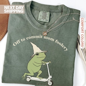 May include: Moss green t-shirt featuring a cartoon frog in a witch hat on a scooter, with the text "Off to commit mom foolery". The shirt has a Comfort Colors label.