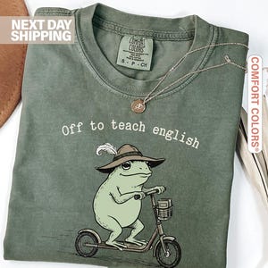 May include: Moss green t-shirt with the text "Off to teach english." The shirt features a cartoon frog wearing a hat and riding a scooter. A gold necklace is draped on the shirt. The shirt is labeled "Comfort Colors."