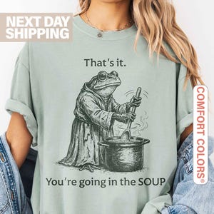 May include: Sage green t-shirt with a frog chef graphic stirring a pot of soup. The text on the shirt reads "That's it. You're going in the SOUP."