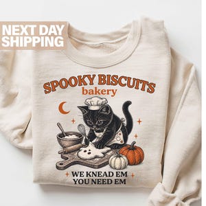 May include: A sand-colored crewneck sweatshirt featuring a graphic of a black cat wearing a chef hat, baking ghost-shaped cookies on a wooden board. The text reads SPOOKY BISCUITS bakery WE KNEAD EM YOU NEED EM, accompanied by a pumpkin.