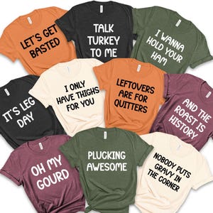 May include: A collection of various colored t-shirts with humorous phrases in white block letters. The shirts include phrases like "Let's Get Basted," "Talk Turkey to Me," and "I Wanna Hold Your Ham."