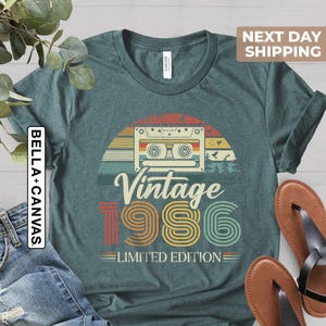 May include: Teal t-shirt with a retro design featuring a cassette tape, a sunset, and the text "Vintage 1986 Limited Edition." The shirt is paired with blue jeans and brown sandals.