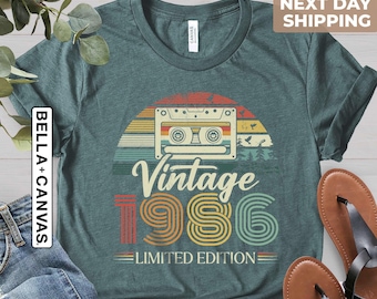 Könnte beinhalten: Türkisfarbenes T-Shirt mit Retro-Design, das eine Kassette, einen Sonnenuntergang und den Text "Vintage 1986 Limited Edition" zeigt. Das Shirt wird mit blauen Jeans und braunen Sandalen kombiniert.