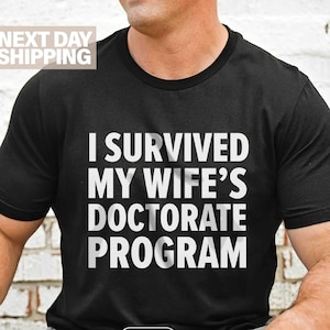Könnte beinhalten: Schwarzes T-Shirt mit weißem Text: "I SURVIVED MY WIFE'S DOCTORATE PROGRAM." Das Shirt hat einen Rundhalsausschnitt und kurze Ärmel. Das Wort "BLACK" ist unten auf dem Shirt aufgedruckt.