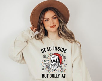 May include: A cream colored sweatshirt with the text "Dead Inside But Jolly AF" in black. A skull wearing a Santa hat and surrounded by Christmas lights is in the center of the text.