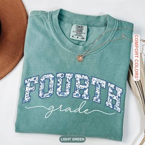 May include: Light green t-shirt with the words "FOURTH grade" in white and blue floral print. The shirt has a crew neck and short sleeves. A silver necklace with a small pendant is on the shirt.
