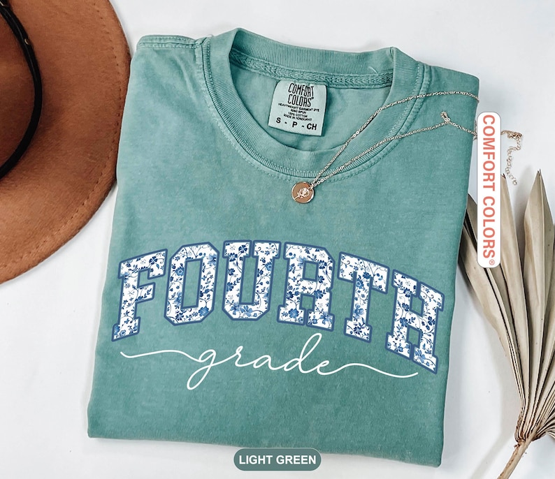 May include: Light green t-shirt with the words "FOURTH grade" in white and blue floral print. The shirt has a crew neck and short sleeves. A silver necklace with a small pendant is on the shirt.
