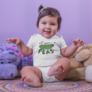 May include: A baby wearing a white onesie with a green pea graphic and the text "Two Peas". The baby is sitting on a colorful rug with a purple hippopotamus and a brown dog plush toy.