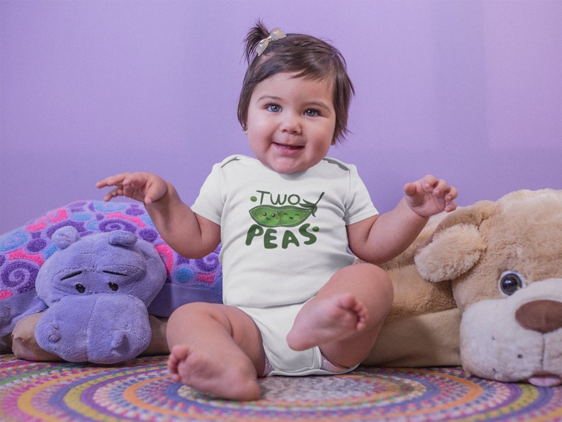May include: A baby wearing a white onesie with a green pea graphic and the text "Two Peas". The baby is sitting on a colorful rug with a purple hippopotamus and a brown dog plush toy.