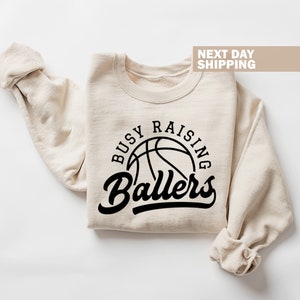 May include: A beige sweatshirt with the text "Busy Raising Ballers" in black lettering. A basketball graphic is inside the circle of text.