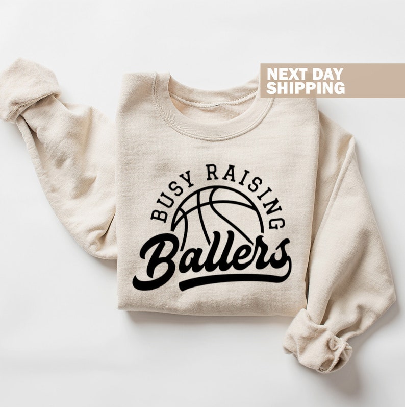 May include: A beige sweatshirt with the text "Busy Raising Ballers" in black lettering. A basketball graphic is inside the circle of text.