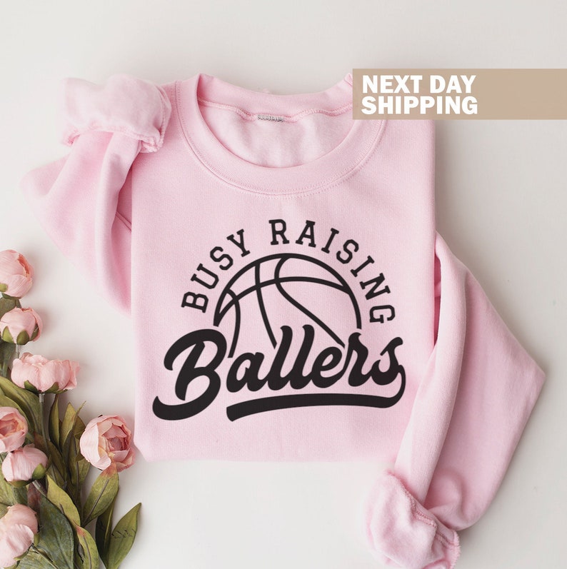 May include: Pink sweatshirt with a black basketball graphic and the text "Busy Raising Ballers".