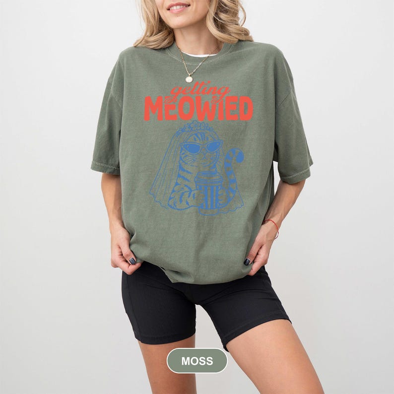 May include: An olive green short-sleeve t-shirt featuring a blue line-art graphic of a cat wearing sunglasses and a bridal veil while holding a drink. The text "getting MEOWIED" is printed in red above the cat illustration. The shirt has a crew neck.