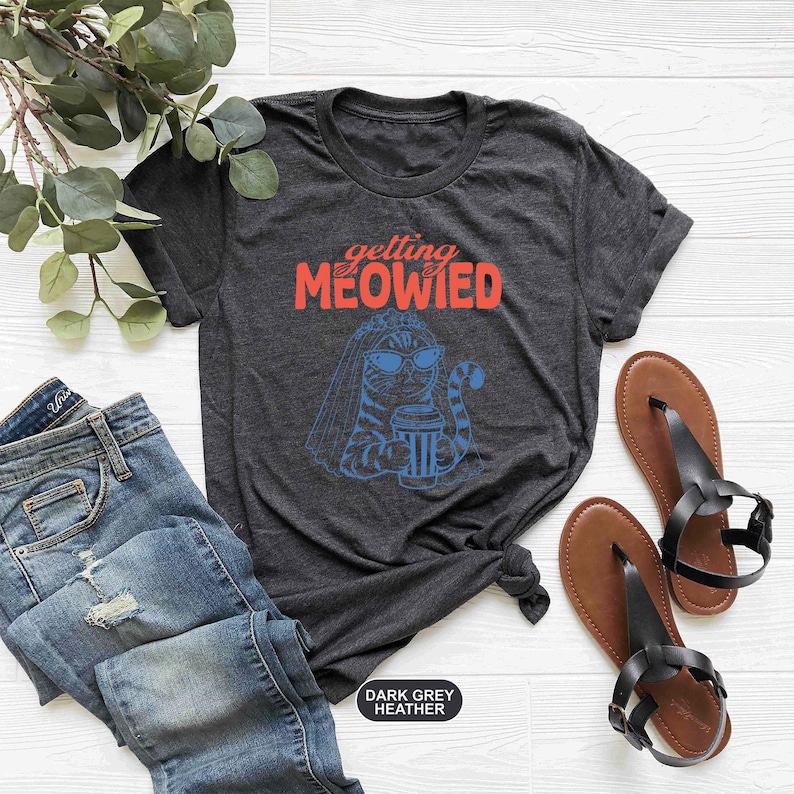 May include: A dark grey heather t-shirt featuring a graphic of a cat wearing a bridal veil and sunglasses while holding a coffee cup. The text above the cat reads "getting MEOWIED" in orange and red lettering. The shirt is short-sleeved with a crew neck.