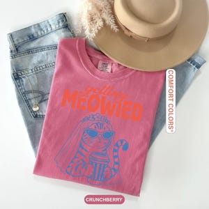 May include: A pink short-sleeved t-shirt featuring the orange text "getting MEOWIED" above a blue graphic of a cat wearing sunglasses and a bridal veil while holding a coffee cup. The shirt is paired with blue denim jeans and a tan wide-brimmed hat.