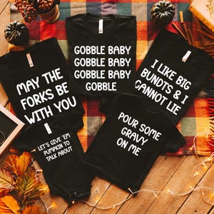 May include: Black t-shirts and a onesie with white text. The shirts say "Gobble Baby", "May the Forks Be With You", "I Like Big Bundts & I Cannot Lie", and "Pour Some Gravy On Me". The onesie says "Let's Give 'Em Pumpkin to Talk About".