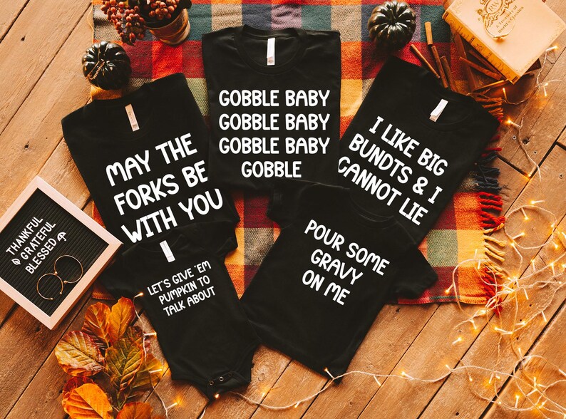 May include: Black t-shirts and a onesie with white text. The shirts say "Gobble Baby", "May the Forks Be With You", "I Like Big Bundts & I Cannot Lie", and "Pour Some Gravy On Me". The onesie says "Let's Give 'Em Pumpkin to Talk About".