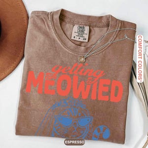May include: A brown short-sleeve t-shirt with the text "getting MEOWIED" in red letters. Below the text is a blue line drawing of a cat wearing sunglasses and a veil. The shirt has a Comfort Colors brand label on the inside collar showing size S.