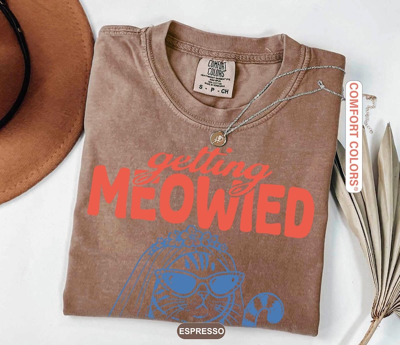 May include: A brown short-sleeve t-shirt with the text "getting MEOWIED" in red letters. Below the text is a blue line drawing of a cat wearing sunglasses and a veil. The shirt has a Comfort Colors brand label on the inside collar showing size S.