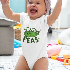 May include: A white baby onesie with a green pea pod design and the text "Two Peas". A baby is wearing the onesie and is sitting on a white rug.