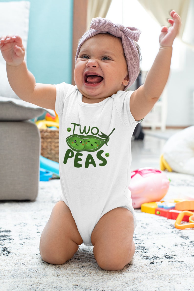 May include: A white baby onesie with a green pea pod design and the text "Two Peas". A baby is wearing the onesie and is sitting on a white rug.