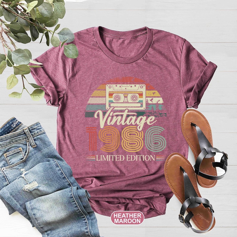 May include: Heather maroon t-shirt with a retro design featuring a cassette tape, the words "Vintage 1986," and "Limited Edition." The shirt is paired with blue jeans and brown sandals.