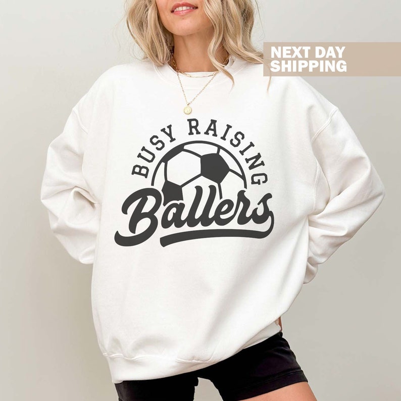 May include: White sweatshirt with black text that reads "Busy Raising Ballers" and a black and white soccer ball graphic.