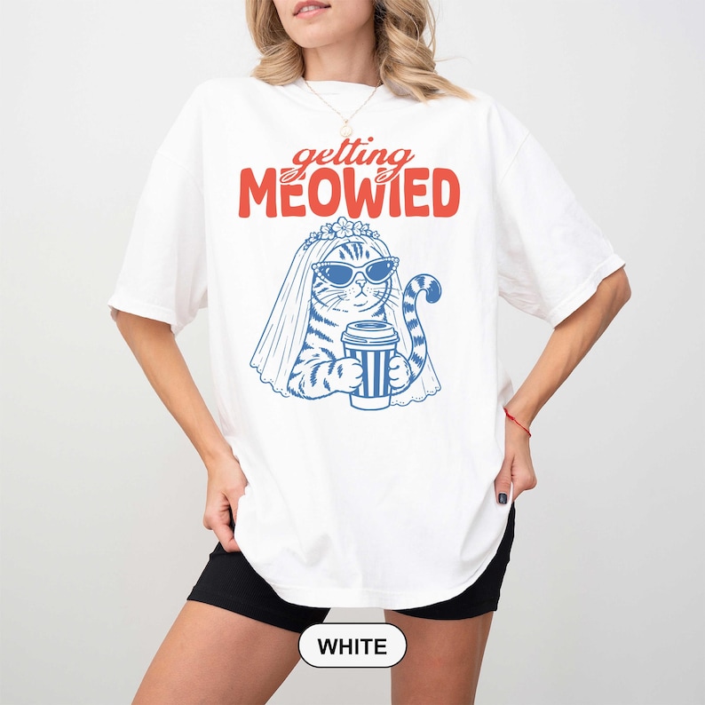 May include: A white t-shirt featuring a graphic of a cat wearing a bridal veil and sunglasses, holding a coffee cup. The text "getting MEOWIED" is printed in red above the cat illustration, which is rendered in blue line art on the front.