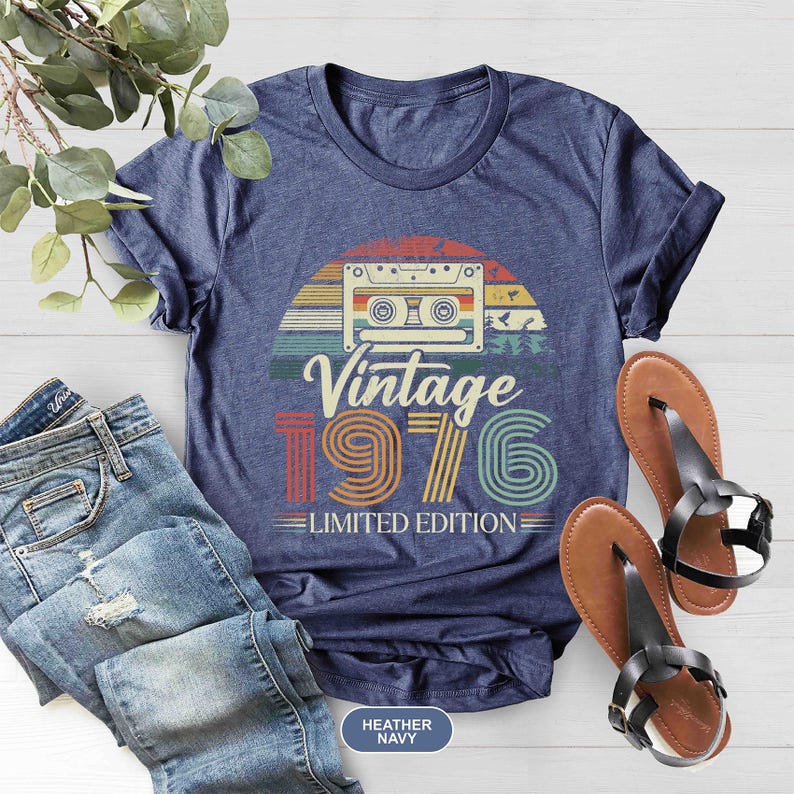 May include: Navy blue t-shirt with a retro design featuring a cassette tape, the words "Vintage 1976," and "Limited Edition." The shirt is paired with blue jeans and brown sandals with black straps.