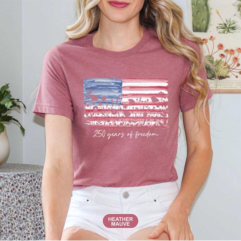 May include: Heather mauve t-shirt featuring a watercolor American flag design. The flag has blue and white stripes with red stripes and floral accents. The text "250 years of freedom" is written below the flag in a script font.