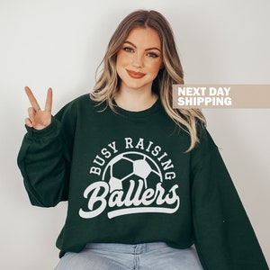 May include: A dark green sweatshirt with a white graphic of a soccer ball and the text "Busy Raising Ballers".