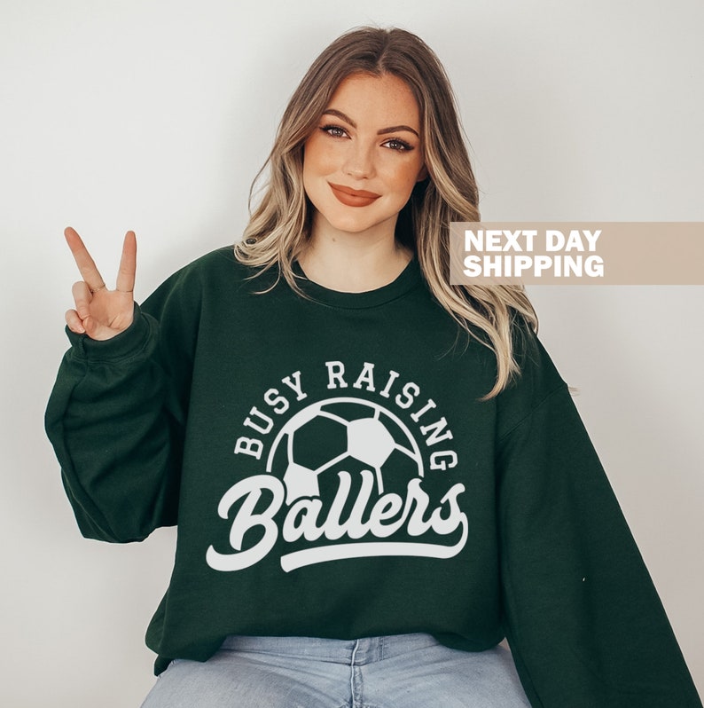 May include: A dark green sweatshirt with a white graphic of a soccer ball and the text "Busy Raising Ballers".