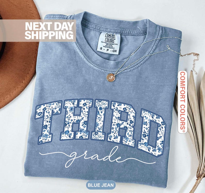 K&ouml;nnte beinhalten: Blaues Jeans-T-Shirt mit den Worten "THIRD grade" in Wei&szlig; mit einem blauen Blumenmuster. Das Shirt hat einen Rundhalsausschnitt und kurze &Auml;rmel. Eine silberne Halskette mit Anh&auml;nger liegt &uuml;ber dem Shirt.