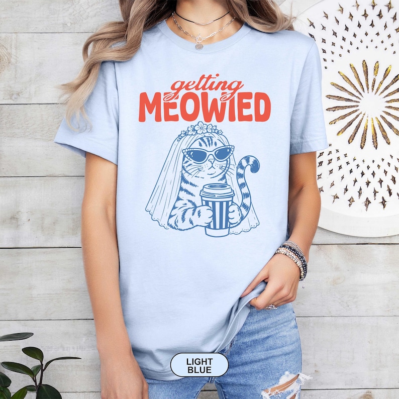 May include: A light blue t-shirt featuring a graphic of a cat wearing sunglasses and a bridal veil while holding a coffee cup. The text above the cat reads "getting MEOWIED" in orange and red lettering. The shirt has short sleeves and a crew neckline.