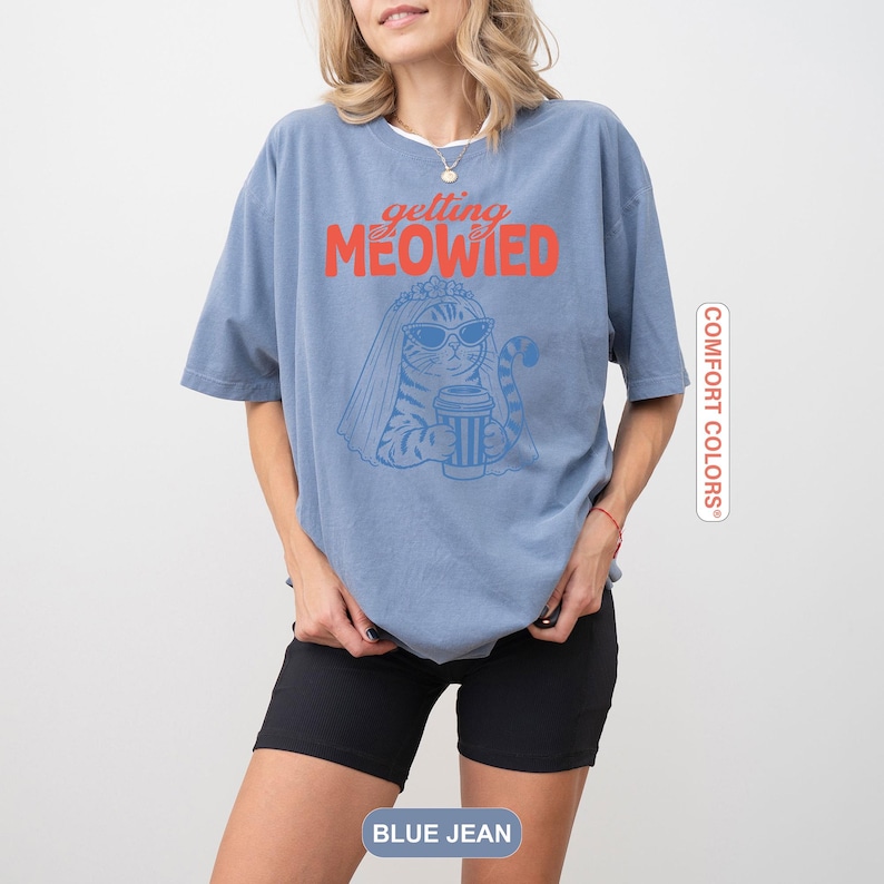 May include: A blue short-sleeve t-shirt featuring a graphic of a cat wearing a bridal veil and sunglasses while holding a coffee cup. The text "getting MEOWIED" is printed in red and blue above the cat illustration. The shirt has a relaxed fit.